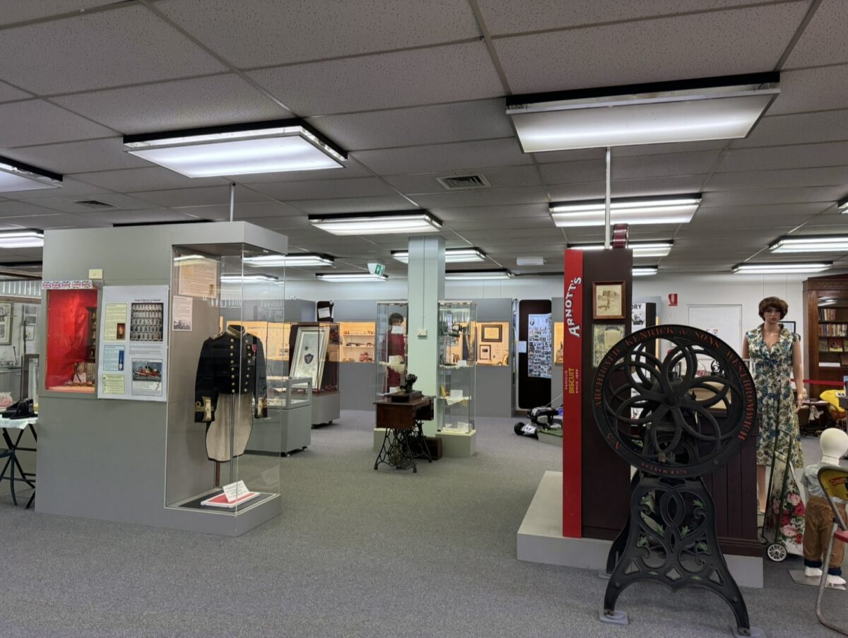 This is an image of the City of Canada Bay Museum. It is a single room, with the grey ceiling, white ceiling LED block lights, and grey carpet visible in the image. There are also several items on display, including an old dark blue army uniform, a old large iron wheel, a mannequin in a green and white floral dress, a small old sewing machine, and a red horizontal sign with the Arnott's logo. There are several other visible display items, but they are harder to identify from the image.